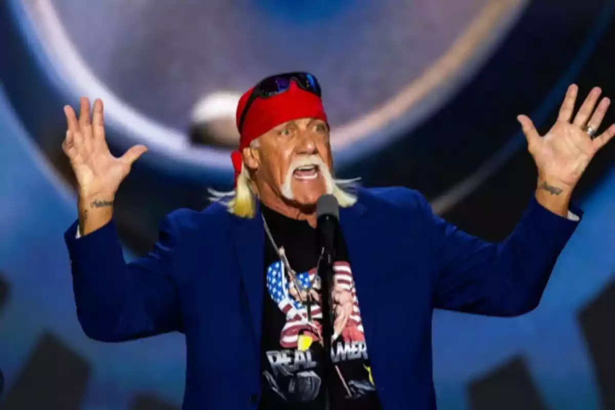 Hombre de cabello largo rubio y bigote blanco usando una bandana roja y gafas de sol en la cabeza, hablando frente a un micrófono con los brazos levantados y vistiendo una camiseta negra y saco azul