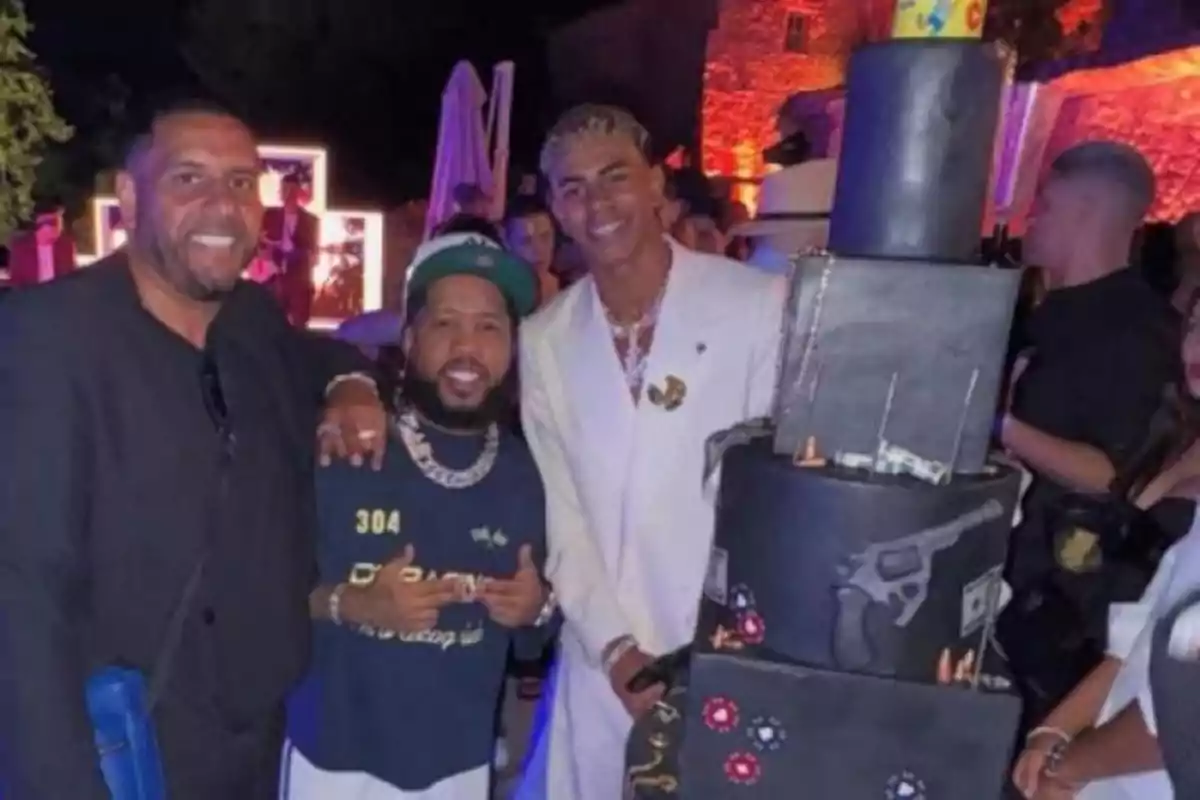 Tres hombres posan sonrientes junto a un pastel de varios niveles decorado con fichas de póker, billetes y la imagen de un revólver en una fiesta nocturna Tres hombres posan sonrientes junto a un pastel de varios niveles decorado con fichas de póker, billetes y la imagen de un revólver en una fiesta nocturna