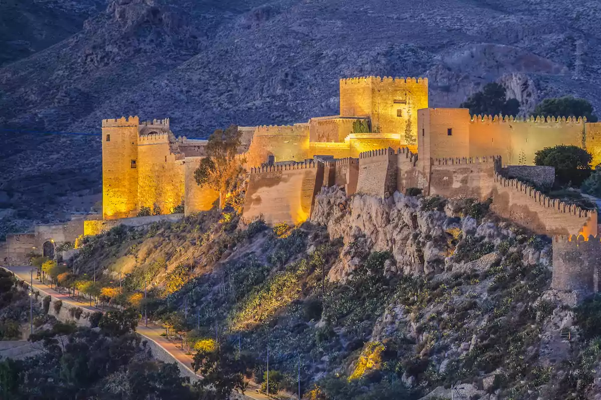 Fortaleza iluminada al atardecer sobre una colina rodeada de vegetación y montañas Fortaleza iluminada al atardecer sobre una colina rodeada de vegetación y montañas