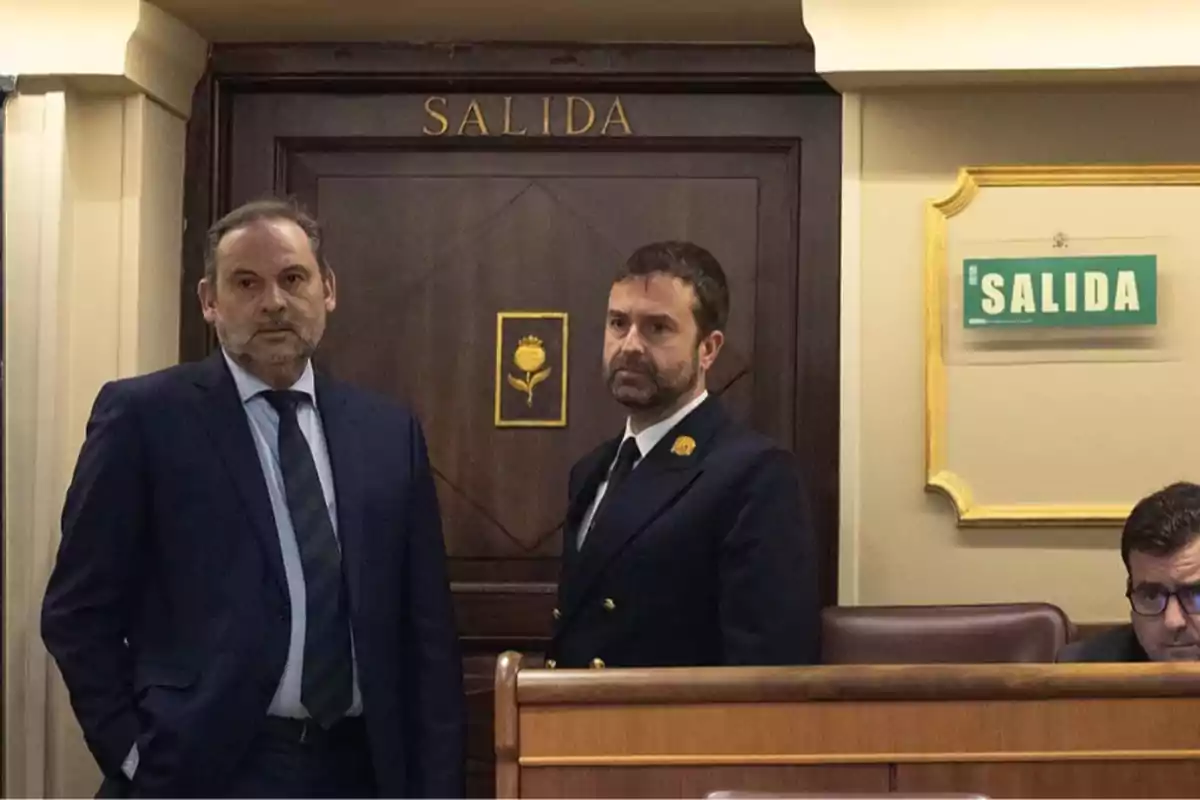 Dos hombres de traje están de pie junto a una puerta de madera con la palabra salida escrita arriba y un letrero verde de salida en la pared Dos hombres de traje están de pie junto a una puerta de madera con la palabra salida escrita arriba y un letrero verde de salida en la pared