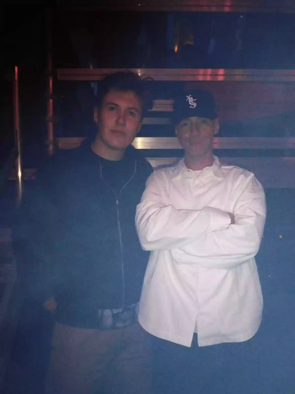 Two people pose together in a dark setting, one wearing a black cap and a white jersey, while the other is dressed in a dark jacket. Two people pose together in a dark setting, one wearing a black cap and a white jersey, while the other is dressed in a dark jacket.
