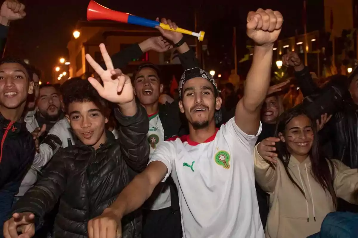 Grupo de jóvenes celebrando con entusiasmo en la calle durante la noche, algunos llevan camisetas de la selección de Marruecos y uno sostiene una vuvuzela Grupo de jóvenes celebrando con entusiasmo en la calle durante la noche, algunos llevan camisetas de la selección de Marruecos y uno sostiene una vuvuzela