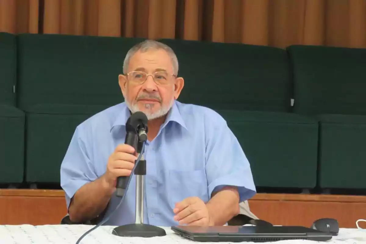 Hombre mayor con barba y gafas hablando por un micrófono mientras está sentado en una mesa con una computadora portátil frente a él Hombre mayor con barba y gafas hablando por un micrófono mientras está sentado en una mesa con una computadora portátil frente a él