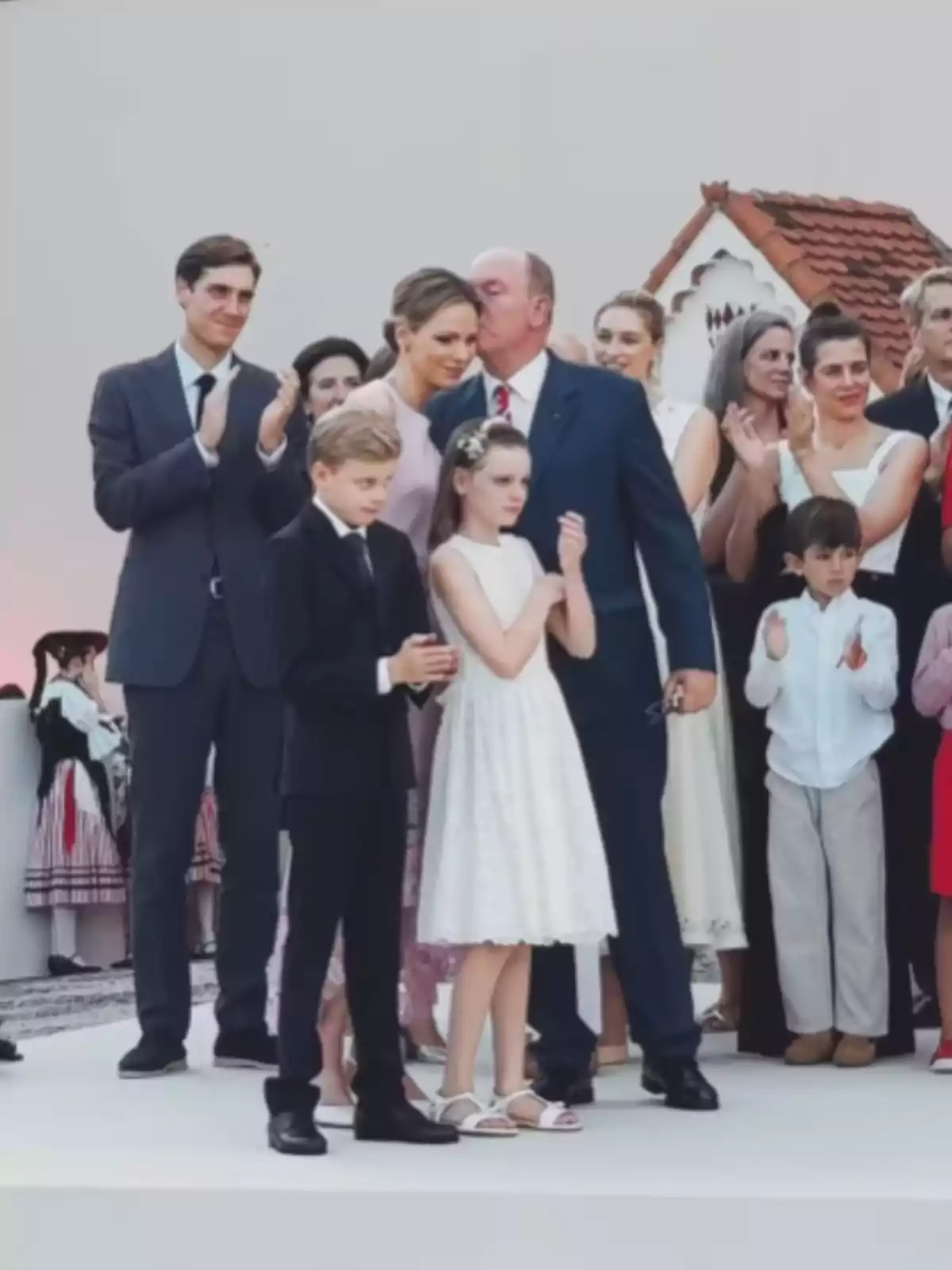 A group of elegantly dressed people gathered on an outdoor stage while some applaud and a man kisses a woman on the head. A group of elegantly dressed people gathered on an outdoor stage while some applaud and a man kisses a woman on the head.