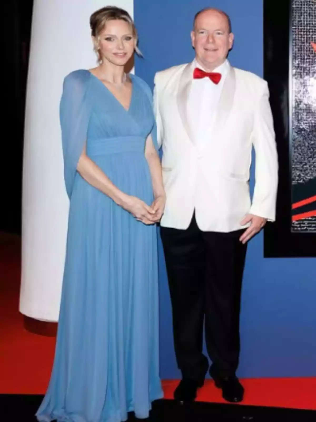 A woman in a long blue dress and a man in a white tuxedo with a red bow tie pose together on a red carpet.