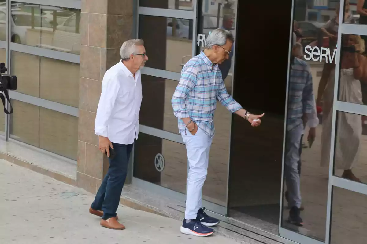Aniceto y Ortega Cano caminan hacia la entrada de un edificio de cristal uno de ellos está abriendo la puerta mientras el otro lo sigue Aniceto y Ortega Cano caminan hacia la entrada de un edificio de cristal uno de ellos está abriendo la puerta mientras el otro lo sigue