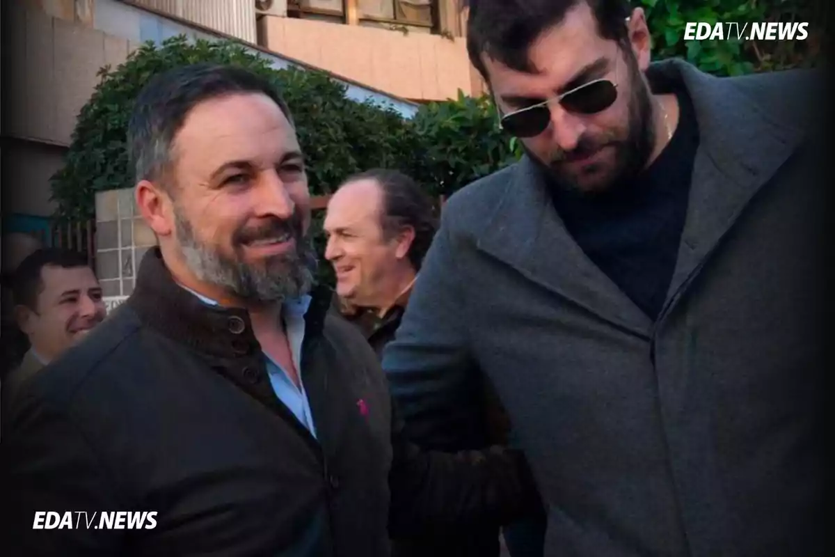 Dos hombres conversan y sonríen al aire libre mientras otras personas se ven al fondo