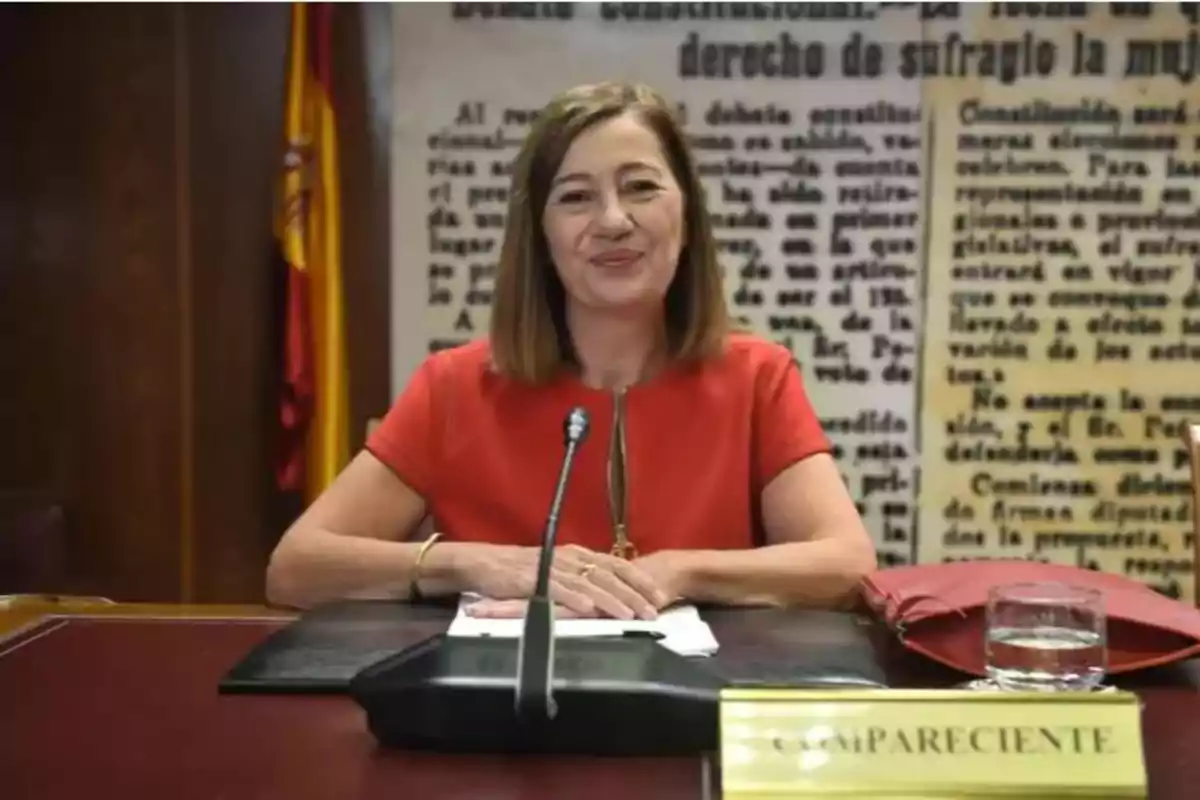 Mujer sentada en una mesa de conferencias con un micrófono frente a ella, una carpeta negra, un vaso de agua y un cartel que dice compareciente, al fondo una bandera de España y un mural con texto antiguo.
