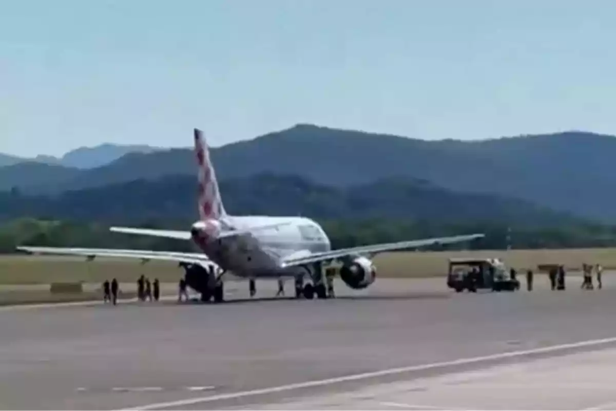 Avión detenido en la pista de un aeropuerto con varias personas y vehículos alrededor y montañas al fondo Avión detenido en la pista de un aeropuerto con varias personas y vehículos alrededor y montañas al fondo