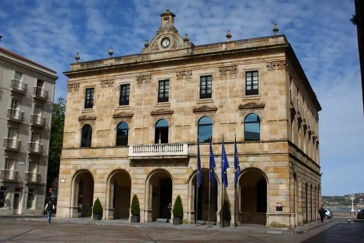 Fachada de un edificio histórico de piedra con reloj en la parte superior y varias banderas azules en la entrada Fachada de un edificio histórico de piedra con reloj en la parte superior y varias banderas azules en la entrada