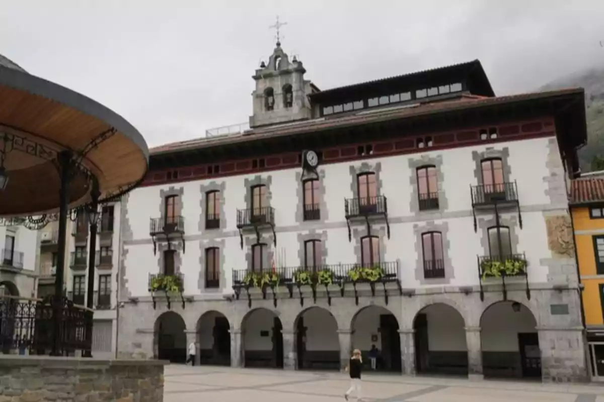 Edificio histórico con arcos en la planta baja, balcones con flores y un reloj en la fachada, situado en una plaza peatonal donde camina una persona Edificio histórico con arcos en la planta baja, balcones con flores y un reloj en la fachada, situado en una plaza peatonal donde camina una persona