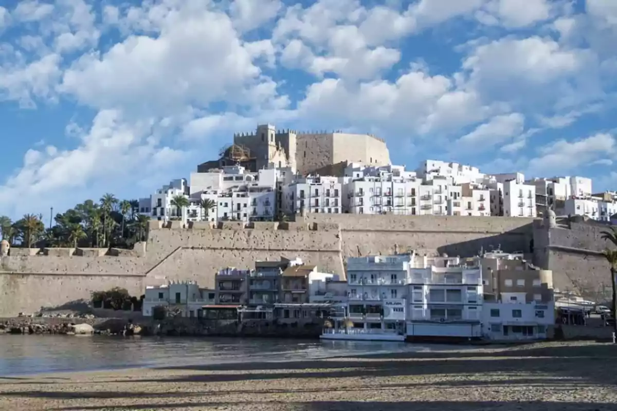 Vista de un castillo medieval en lo alto de una colina rodeado de casas blancas y murallas junto al mar bajo un cielo azul con nubes
