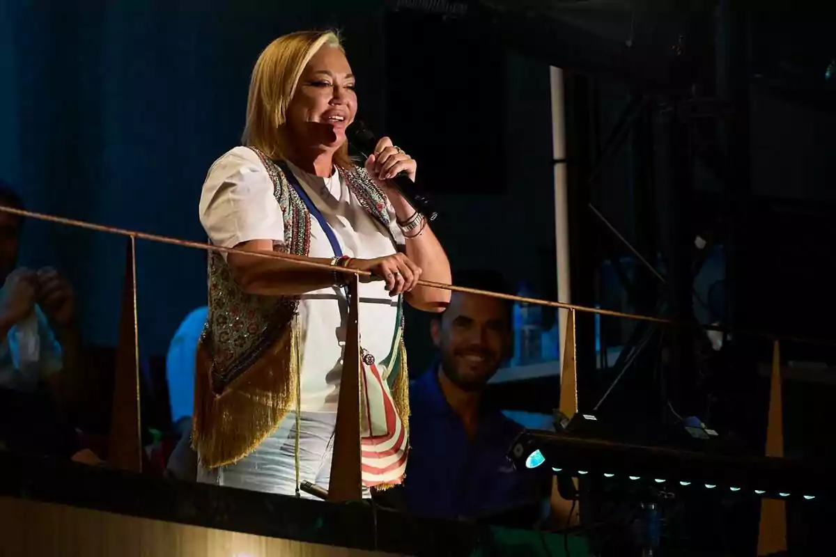 Belén Esteban sonriente con micrófono en la mano vestida con camiseta blanca y chaleco colorido de pie en un balcón durante un evento nocturno. Belén Esteban sonriente con micrófono en la mano vestida con camiseta blanca y chaleco colorido de pie en un balcón durante un evento nocturno.