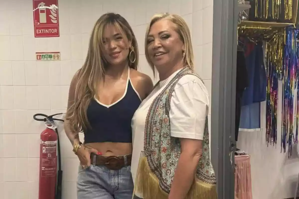 Dos mujeres, que son Karol G y Belén Esteban, posan sonrientes en una habitación decorada con cortinas de colores y un extintor en la pared. Dos mujeres, que son Karol G y Belén Esteban, posan sonrientes en una habitación decorada con cortinas de colores y un extintor en la pared.
