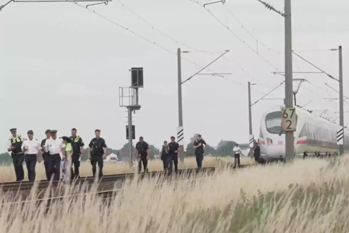 Varios policías caminan junto a las vías del tren mientras un tren blanco se encuentra detenido al fondo en un entorno rural.