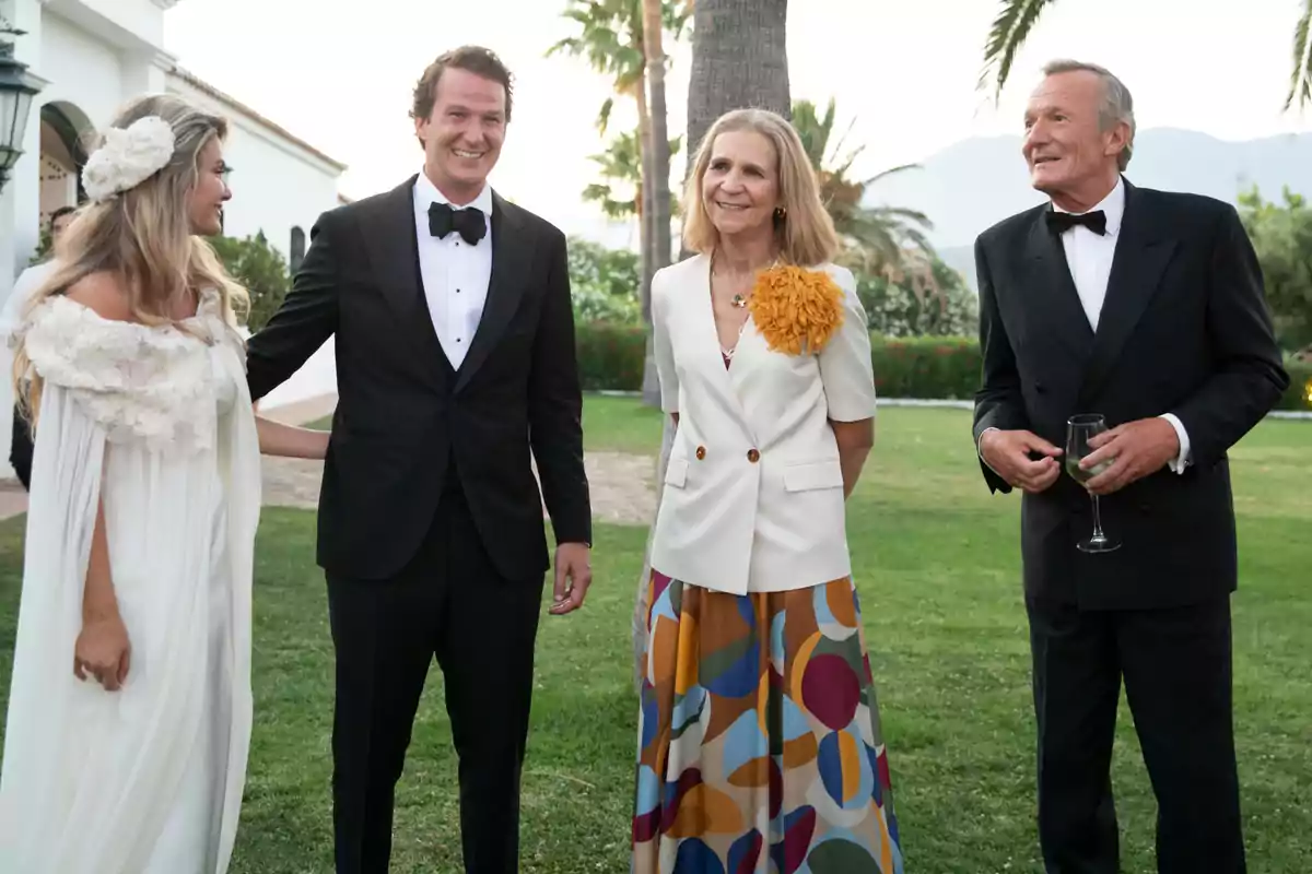 Cristina Serrano, Luis Astolfi Jr, Infanta Elena, and Luis Astolfi, elegantly dressed, pose and smile outdoors in a garden, with palm trees and mountains in the background.