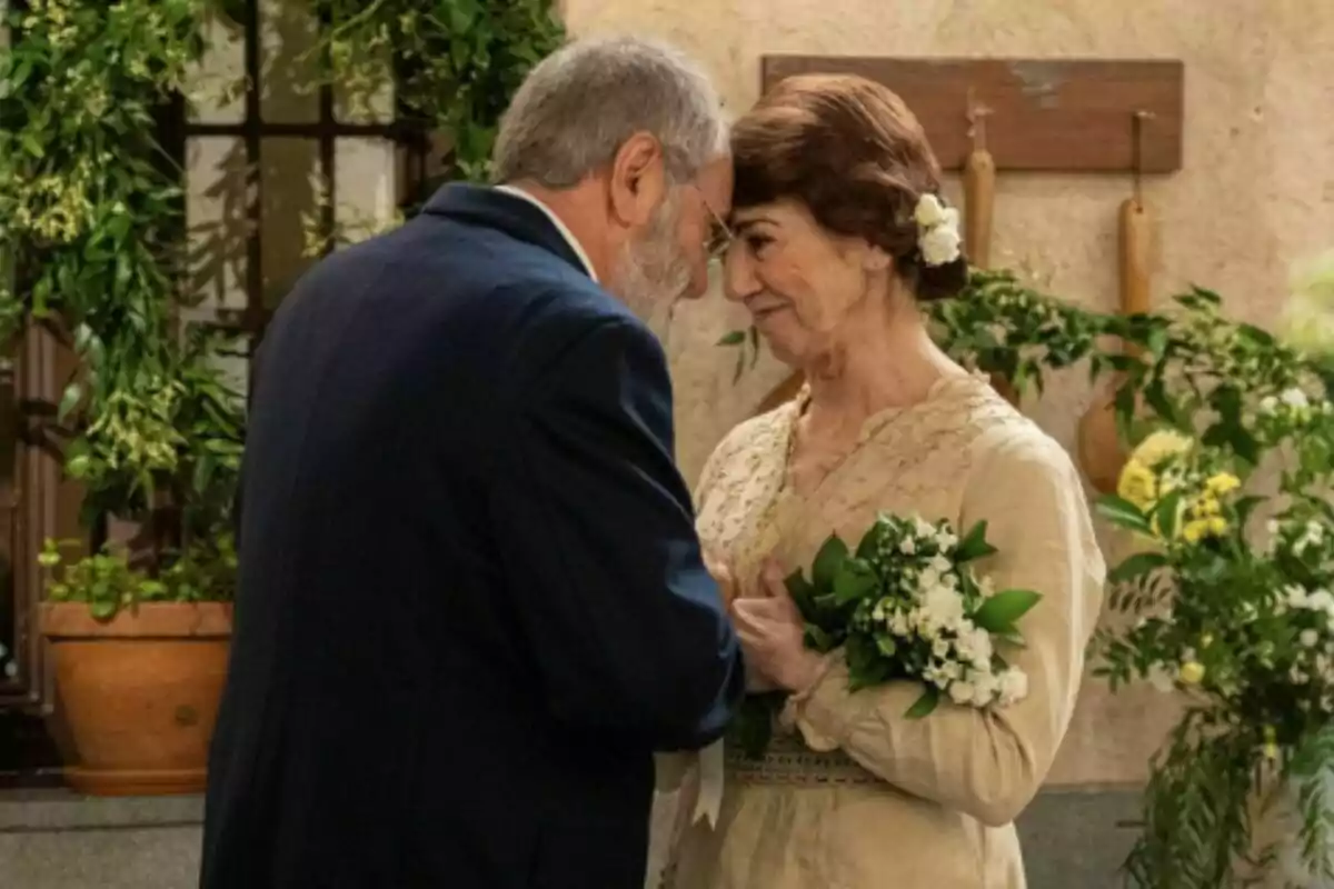 Pareja de personas mayores, formada por Rómulo y Emilia en 'La Promesa', vestidas de manera elegante se miran con ternura y se tocan la frente mientras la mujer sostiene un ramo de flores blancas rodeados de plantas y decoración natural. Pareja de personas mayores, formada por Rómulo y Emilia en 'La Promesa', vestidas de manera elegante se miran con ternura y se tocan la frente mientras la mujer sostiene un ramo de flores blancas rodeados de plantas y decoración natural.