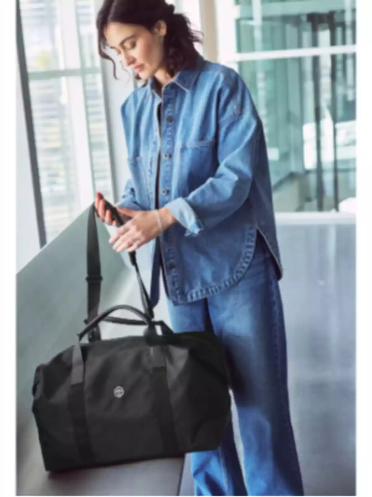 Mujer con camisa y pantalón de mezclilla sosteniendo una bolsa de viaje negra en un entorno moderno y luminoso Mujer con camisa y pantalón de mezclilla sosteniendo una bolsa de viaje negra en un entorno moderno y luminoso