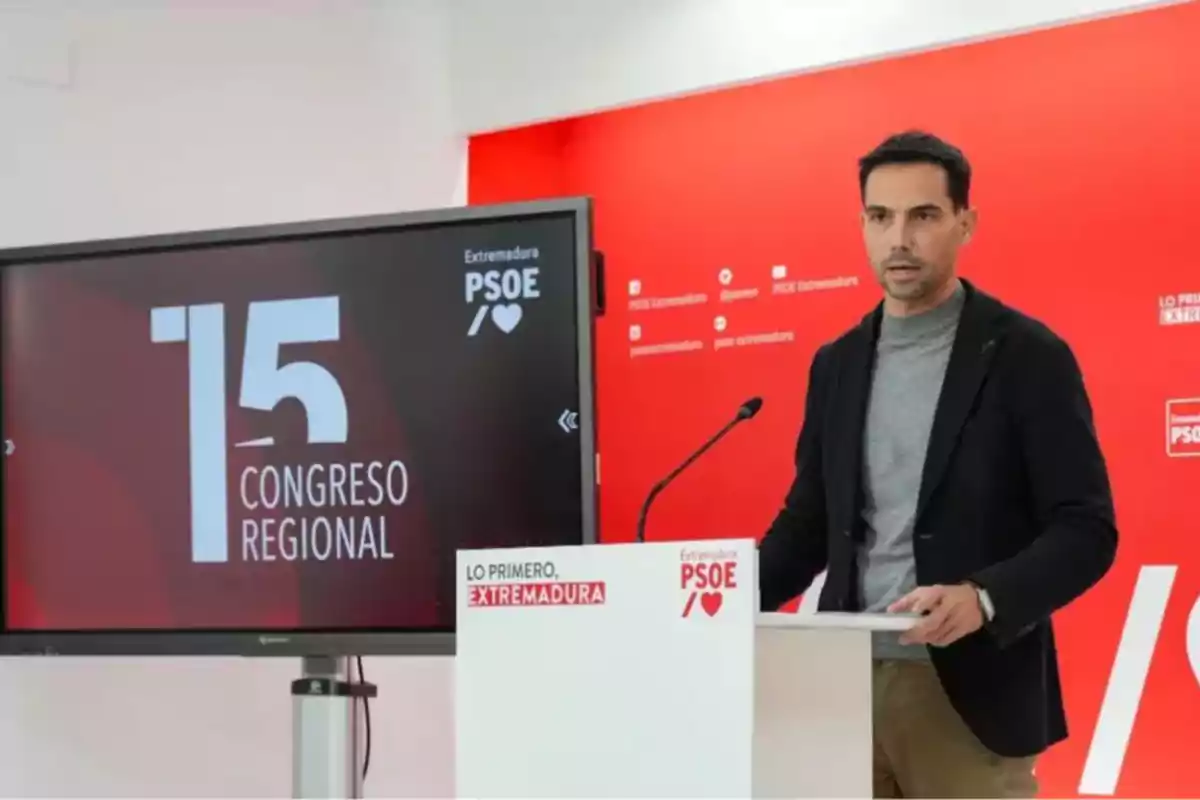 Un hombre de pie en un atril con fondo rojo y una pantalla que muestra el texto 15 Congreso Regional del PSOE de Extremadura Un hombre de pie en un atril con fondo rojo y una pantalla que muestra el texto 15 Congreso Regional del PSOE de Extremadura