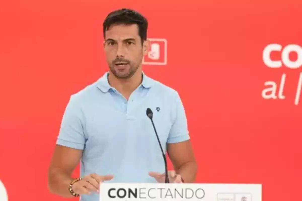 Hombre de cabello corto y barba hablando en un podio con fondo rojo y la palabra conectando escrita en el atril