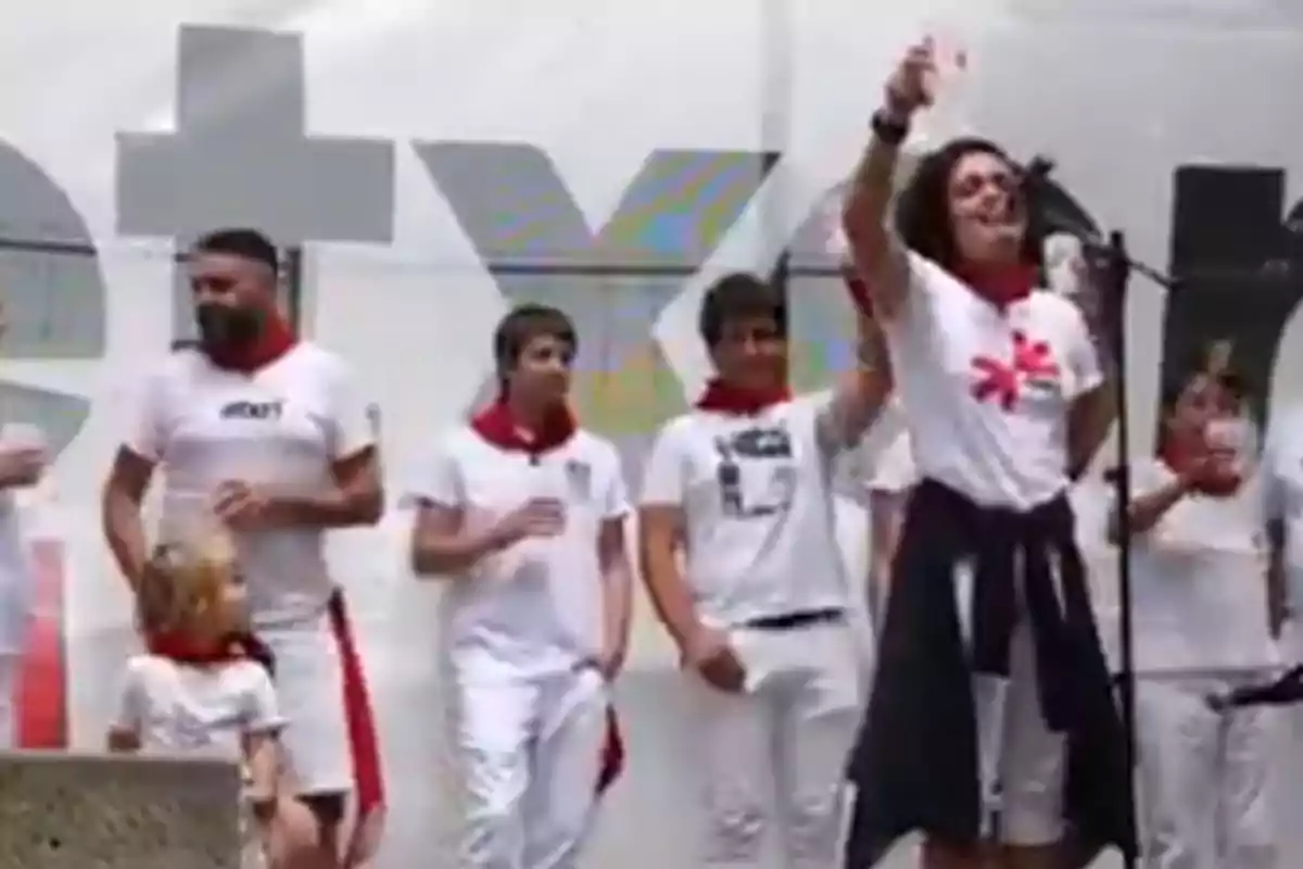 Grupo de personas vestidas de blanco con pañuelos rojos celebrando en un escenario mientras una persona habla o canta al micrófono Grupo de personas vestidas de blanco con pañuelos rojos celebrando en un escenario mientras una persona habla o canta al micrófono