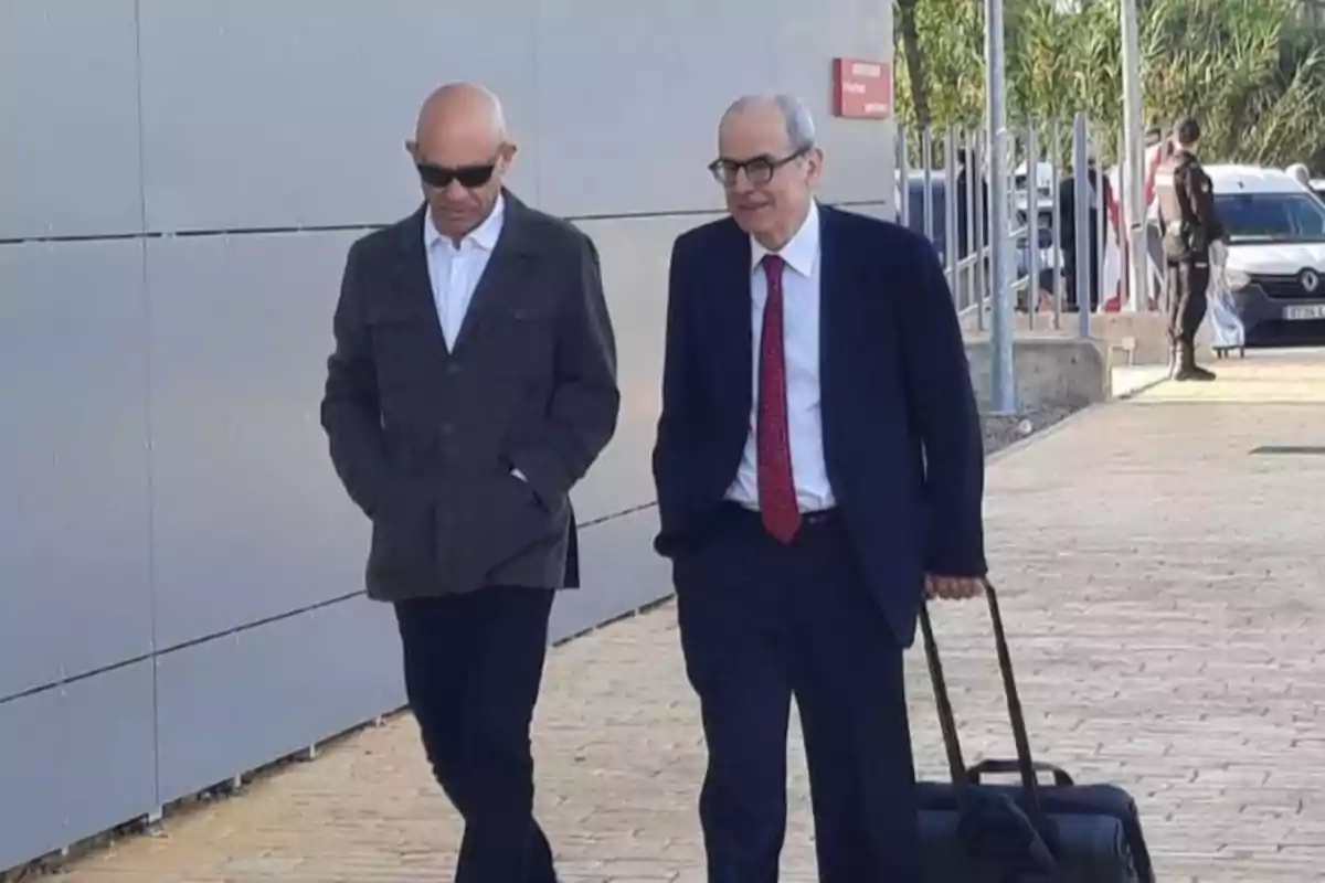 Dos hombres caminan juntos por una acera uno de ellos lleva una maleta de ruedas y ambos visten ropa formal