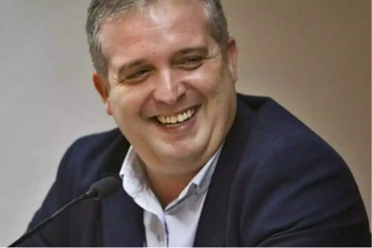 Hombre de cabello corto y canoso sonriendo mientras habla frente a un micrófono vestido con saco azul y camisa blanca Hombre de cabello corto y canoso sonriendo mientras habla frente a un micrófono vestido con saco azul y camisa blanca