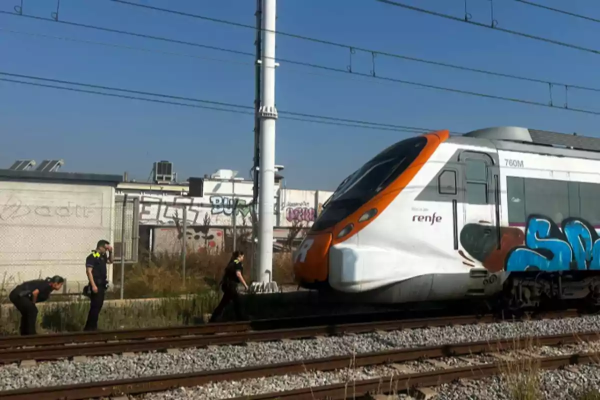 Un tren detenido en las vías mientras tres policías inspeccionan la zona junto a las vías y una pared con grafitis al fondo