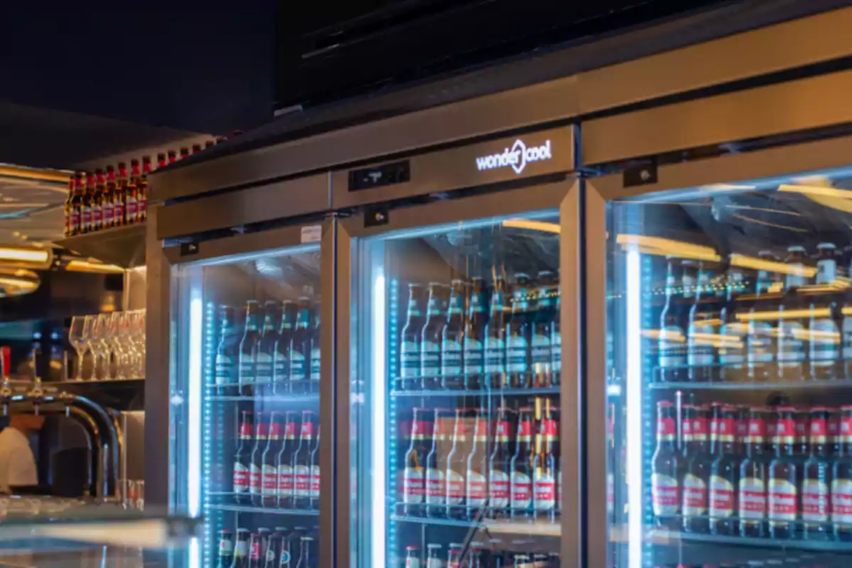 Refrigerador de bar con puertas de vidrio lleno de botellas de cerveza y copas de cristal al lado
