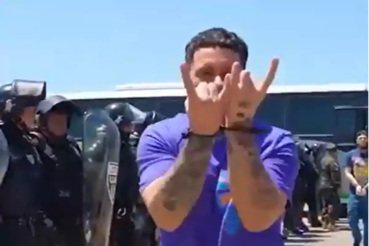 Hombre esposado con camiseta morada haciendo un gesto con las manos mientras policías antidisturbios están alineados al fondo Hombre esposado con camiseta morada haciendo un gesto con las manos mientras policías antidisturbios están alineados al fondo