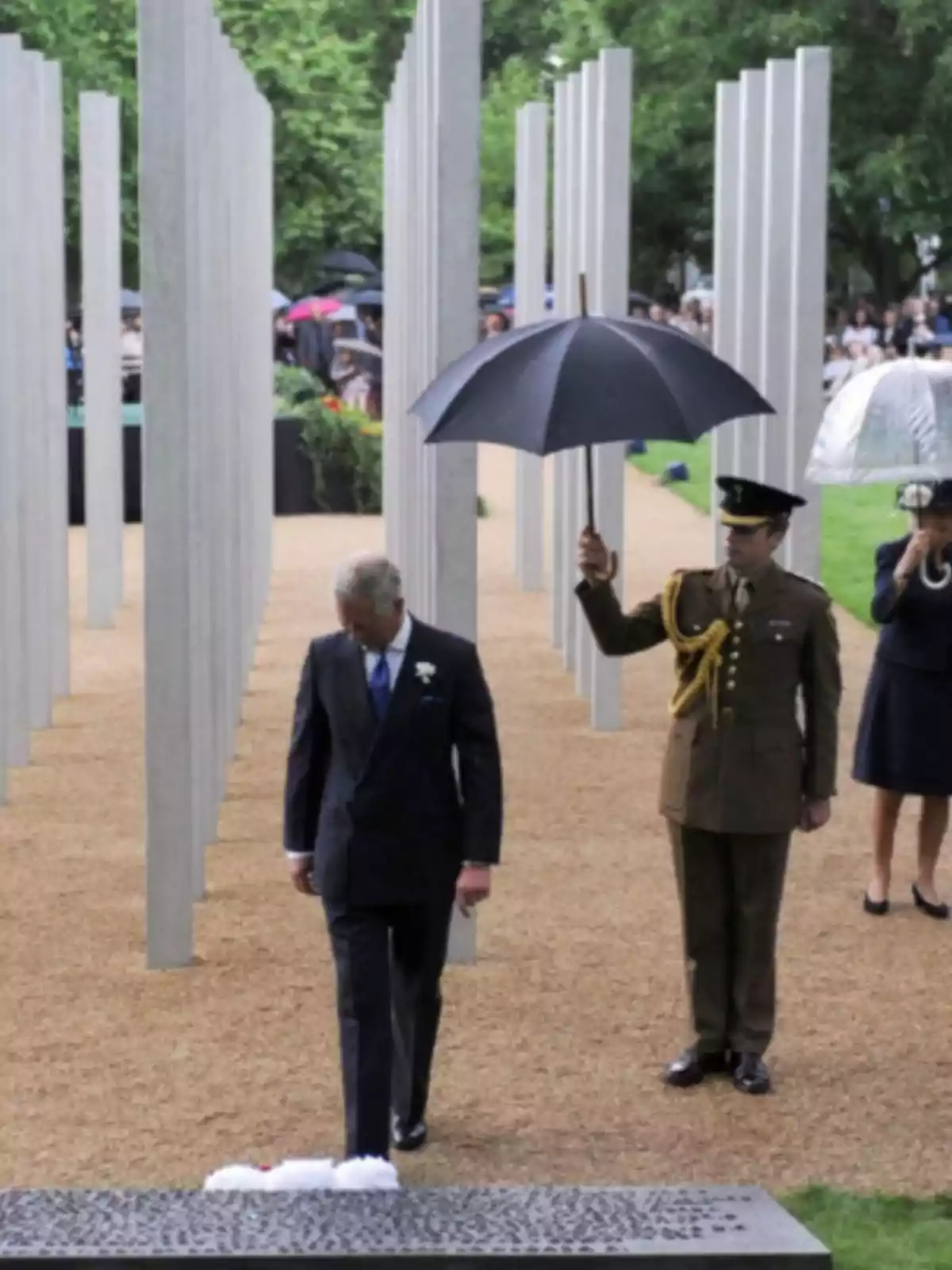 Un hombre de traje camina cabizbajo mientras un militar sostiene un paraguas sobre él en un monumento con columnas verticales y personas al fondo Un hombre de traje camina cabizbajo mientras un militar sostiene un paraguas sobre él en un monumento con columnas verticales y personas al fondo