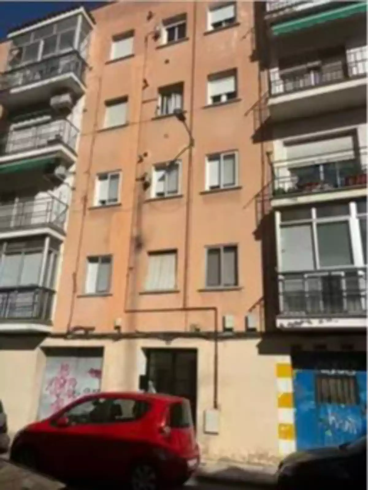 Fachada de un edificio de apartamentos de varios pisos con paredes color salmón y ventanas blancas, en la planta baja hay un coche rojo estacionado y una puerta azul con grafitis Fachada de un edificio de apartamentos de varios pisos con paredes color salmón y ventanas blancas, en la planta baja hay un coche rojo estacionado y una puerta azul con grafitis