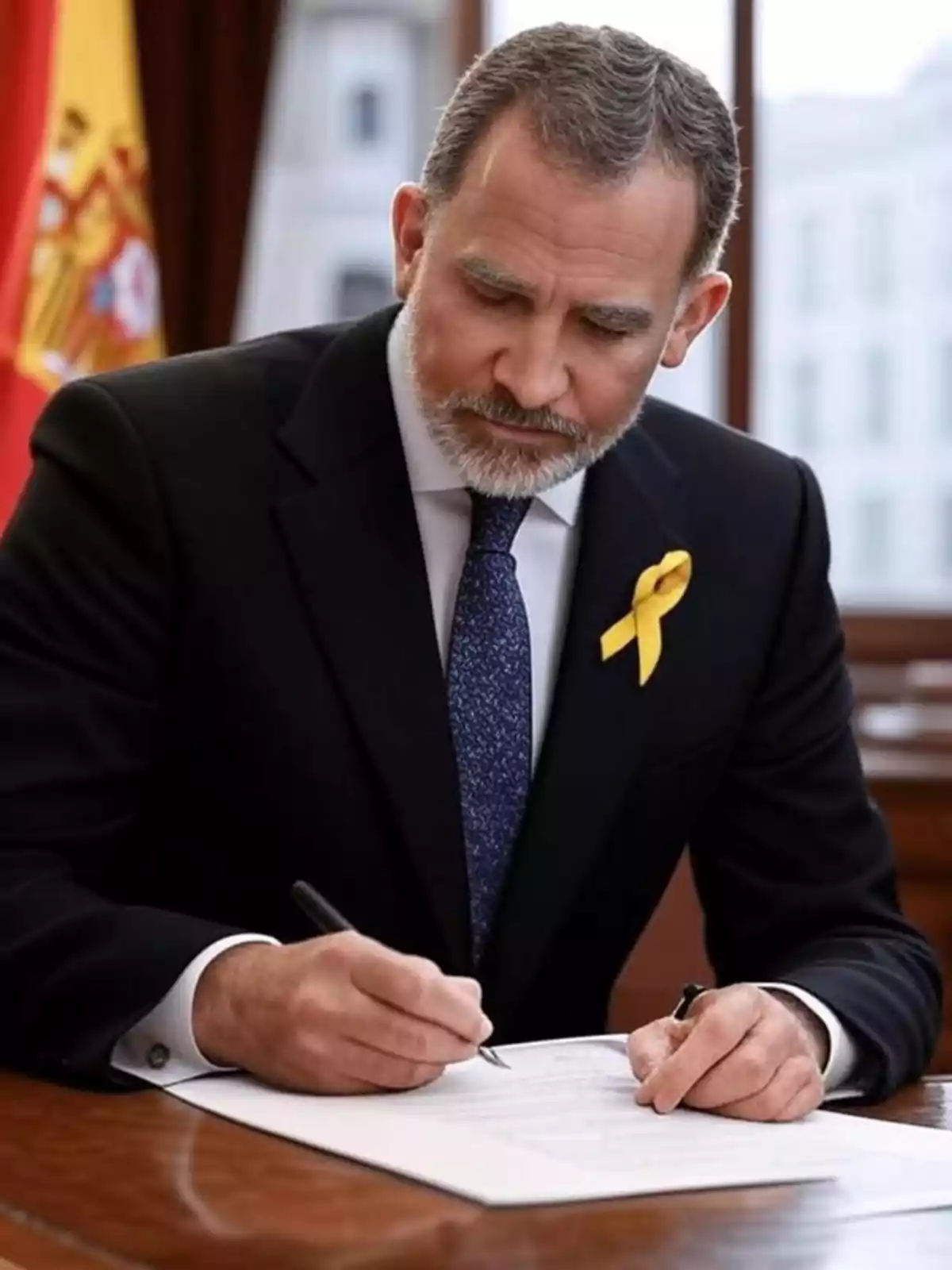 Hombre de traje oscuro con corbata azul y lazo amarillo en la solapa firmando un documento en una oficina con una bandera de España al fondo Hombre de traje oscuro con corbata azul y lazo amarillo en la solapa firmando un documento en una oficina con una bandera de España al fondo