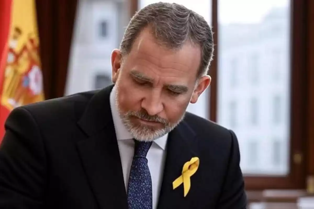 Hombre de cabello y barba canosa con traje oscuro y corbata azul, lleva un lazo amarillo en la solapa y está sentado frente a una ventana con una bandera de España al fondo