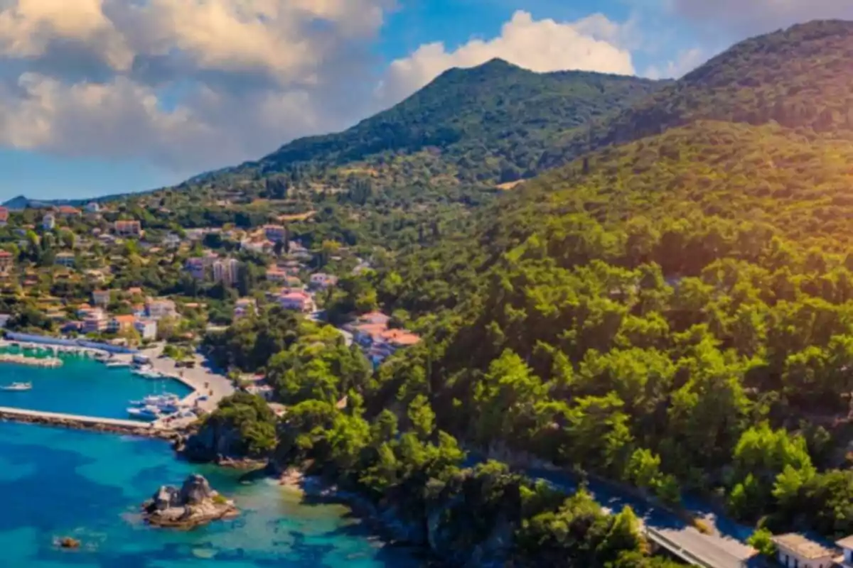 Vista panorámica de un pueblo costero rodeado de colinas verdes y mar azul bajo un cielo parcialmente nublado Vista panorámica de un pueblo costero rodeado de colinas verdes y mar azul bajo un cielo parcialmente nublado