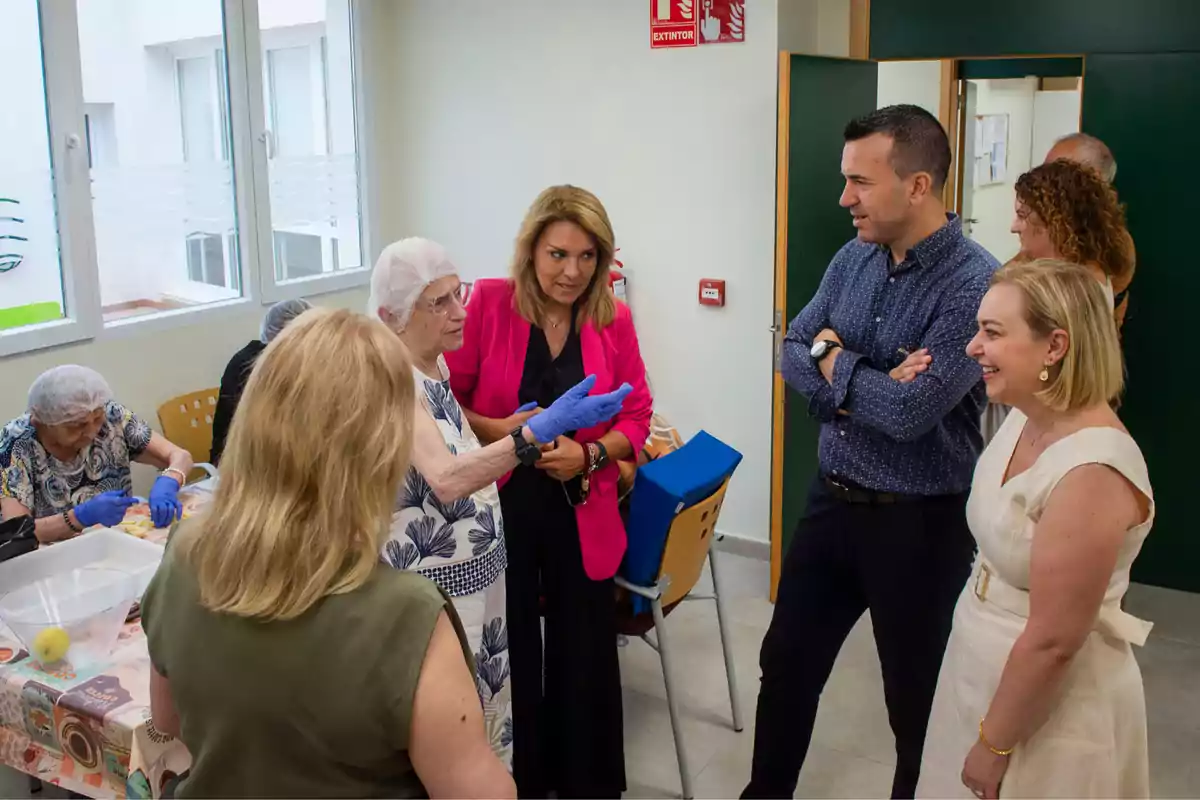 Varias personas conversan y observan a un grupo de mujeres mayores que participan en una actividad en una sala iluminada con ventanas grandes