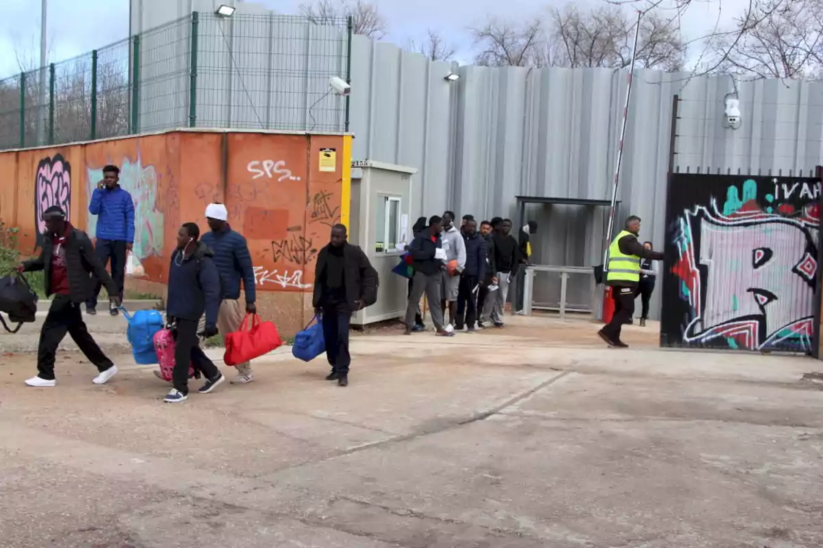 Un grupo de personas camina con maletas y bolsas saliendo de un recinto cercado con paredes de metal y grafitis, mientras un hombre con chaleco reflectante supervisa la salida.