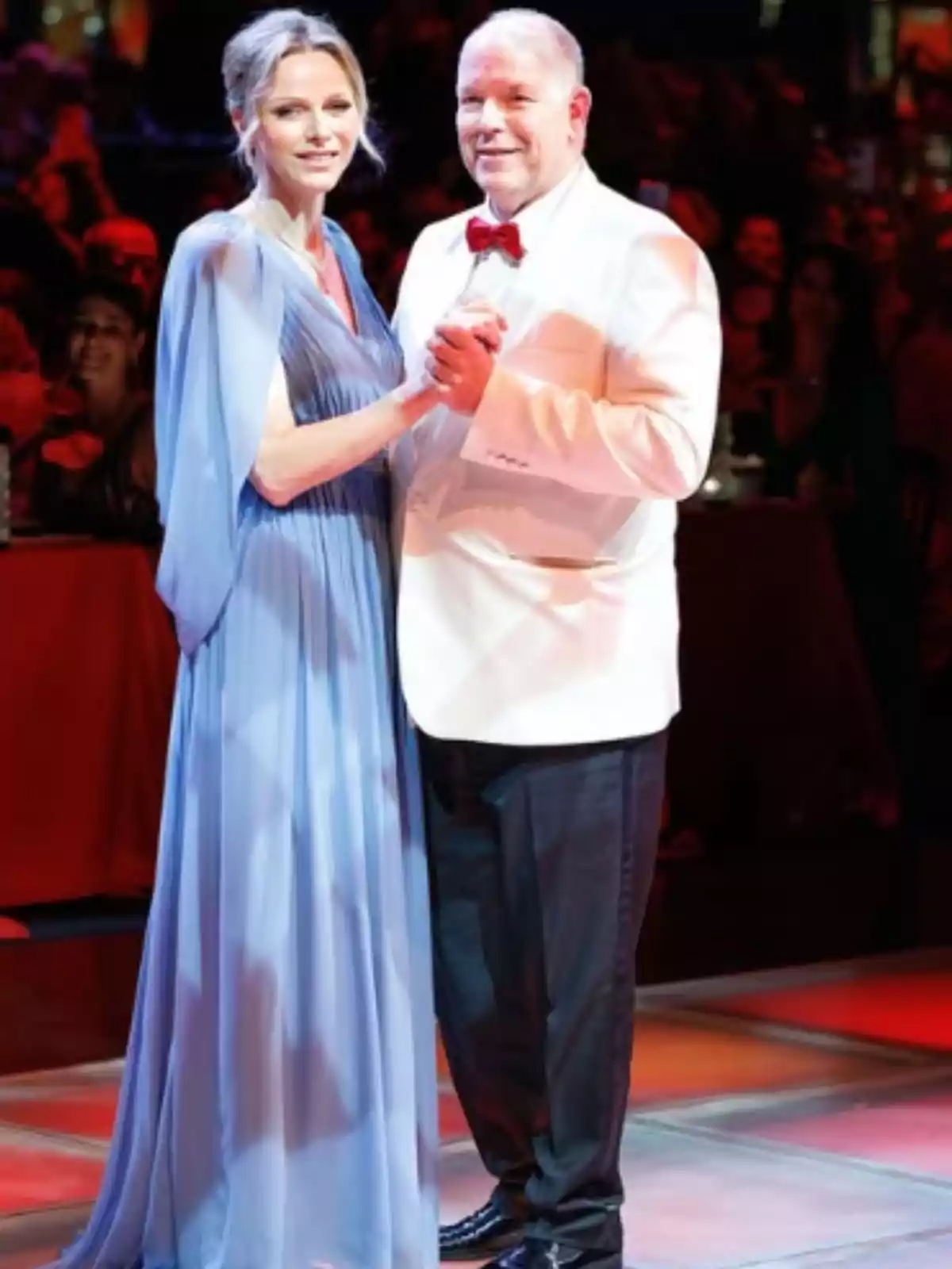 An elegantly dressed couple dances at a formal event with warm lights and an audience in the background.
