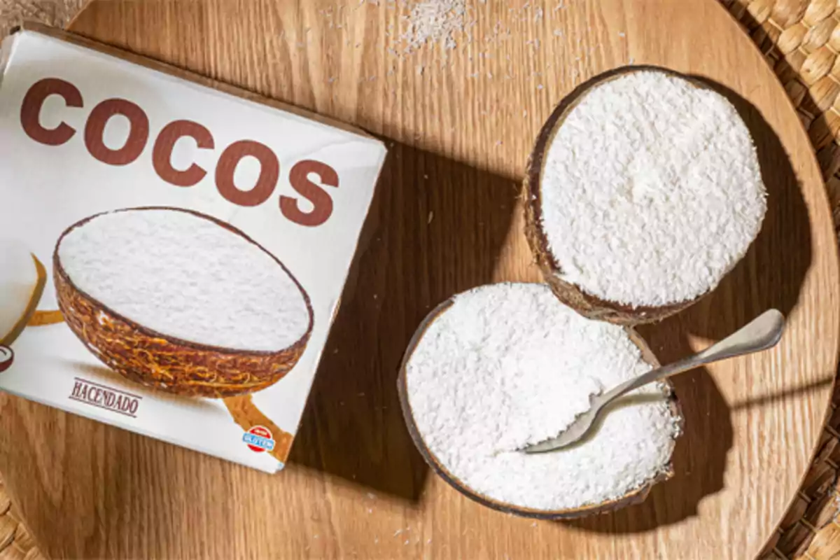 Caja de coco rallado de la marca Hacendado junto a dos mitades de coco rellenas de coco rallado y una cuchara sobre una bandeja de madera Caja de coco rallado de la marca Hacendado junto a dos mitades de coco rellenas de coco rallado y una cuchara sobre una bandeja de madera