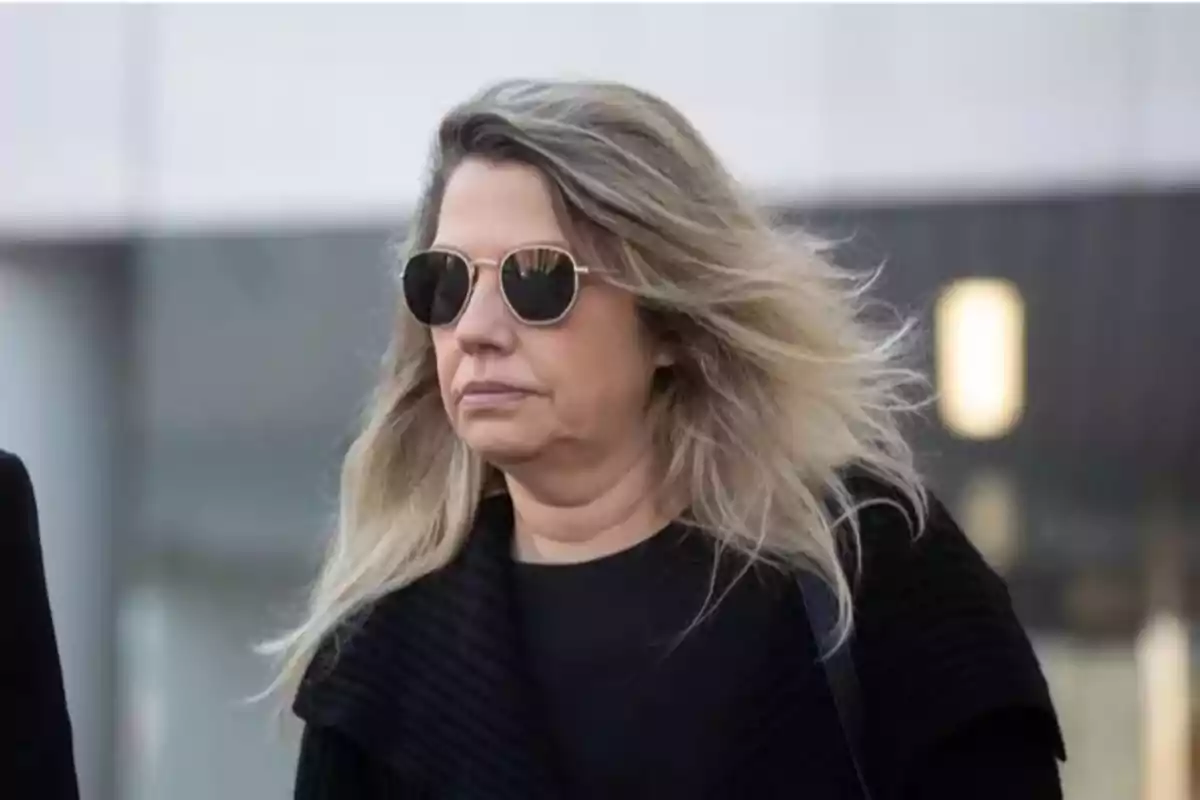 Mujer de cabello rubio y largo usando gafas de sol y ropa oscura caminando al aire libre