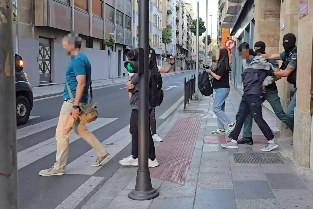 Un hombre es arrestado por dos agentes encapuchados en una acera mientras otras personas caminan cerca en una calle urbana. Un hombre es arrestado por dos agentes encapuchados en una acera mientras otras personas caminan cerca en una calle urbana.