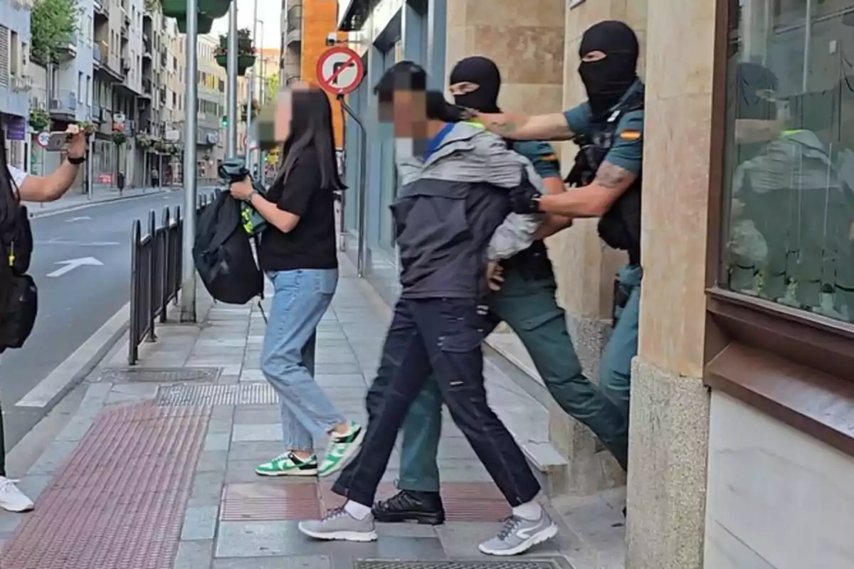 Un agente con pasamontañas escolta a una persona esposada por la acera mientras otra persona observa y graba con un teléfono móvil en una calle urbana