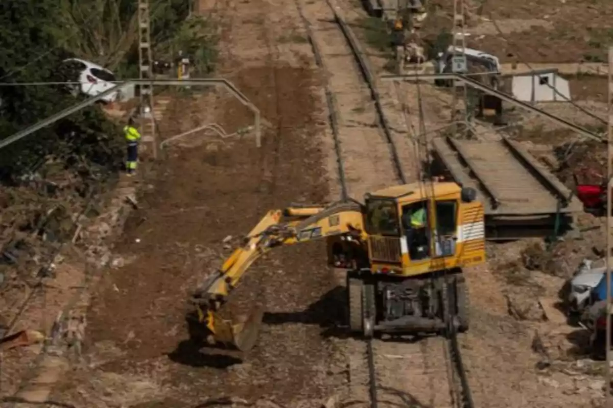 Excavadora trabajando sobre unas vías de tren cubiertas de tierra mientras un trabajador con chaleco reflectante observa y varios coches están fuera de la carretera Excavadora trabajando sobre unas vías de tren cubiertas de tierra mientras un trabajador con chaleco reflectante observa y varios coches están fuera de la carretera