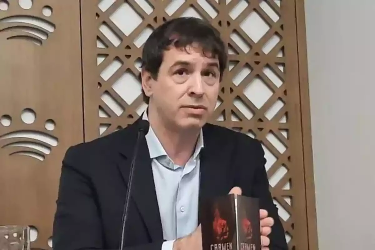 Hombre de cabello oscuro con saco y camisa clara sentado frente a un micrófono sosteniendo un libro titulado Carmen con una portada oscura y fondo de panel decorativo de madera. Hombre de cabello oscuro con saco y camisa clara sentado frente a un micrófono sosteniendo un libro titulado Carmen con una portada oscura y fondo de panel decorativo de madera.