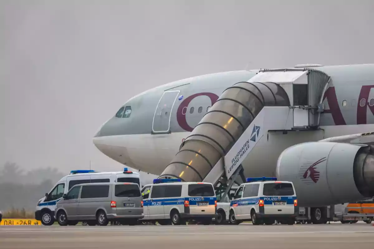 Un avión de Qatar Airways estacionado en la pista junto a varias camionetas de la policía y una escalera de embarque en un aeropuerto bajo cielo nublado. Un avión de Qatar Airways estacionado en la pista junto a varias camionetas de la policía y una escalera de embarque en un aeropuerto bajo cielo nublado.