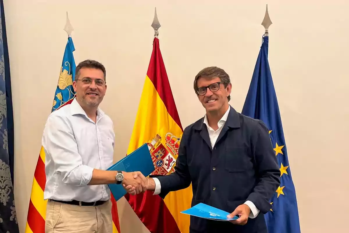 Dos hombres sonríen y se dan la mano mientras sostienen carpetas azules frente a las banderas de la Comunidad Valenciana, España y la Unión Europea