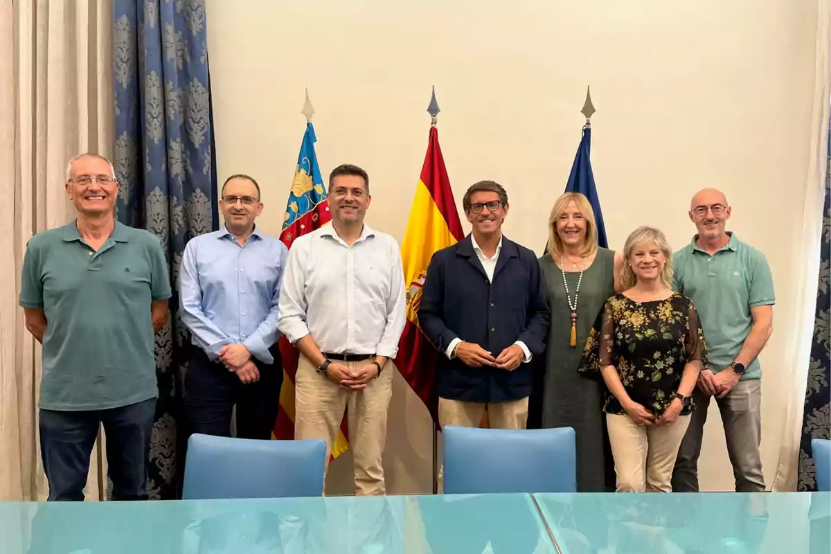Siete personas posan sonrientes frente a tres banderas en una sala de reuniones con cortinas y sillas azules Siete personas posan sonrientes frente a tres banderas en una sala de reuniones con cortinas y sillas azules
