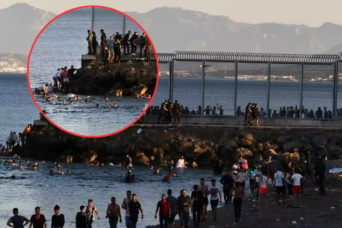 Un grupo numeroso de personas cruza nadando hacia la costa mientras otras caminan por la playa y varias más son observadas por autoridades cerca de una valla fronteriza junto al mar, con montañas al fondo.