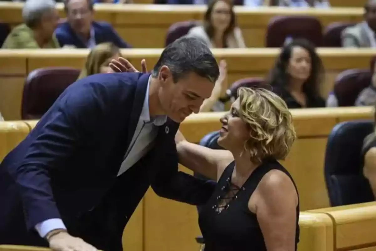 Un hombre y una mujer se saludan sonrientes en un recinto con asientos tipo auditorio y varias personas al fondo.