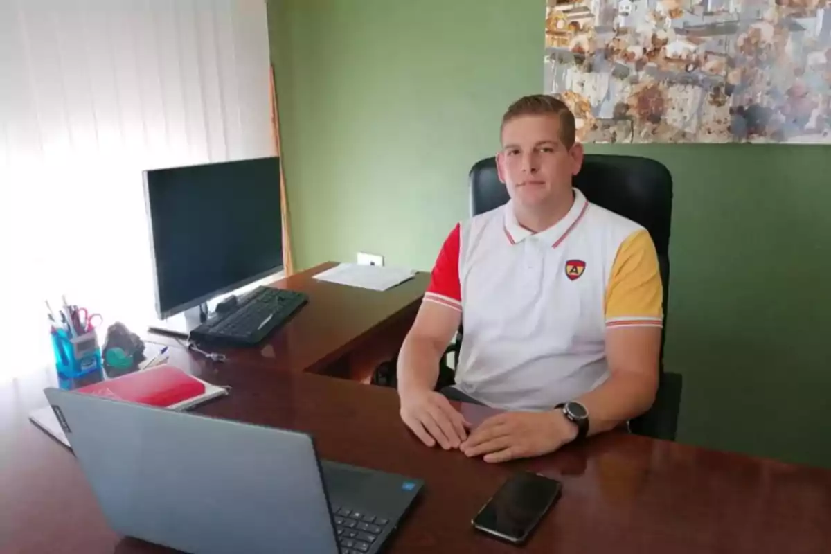 Hombre joven sentado en un escritorio de oficina con una laptop, un monitor, útiles de oficina y un teléfono móvil, vistiendo una camiseta blanca con detalles amarillos y rojos, fondo de pared verde y cuadro decorativo.